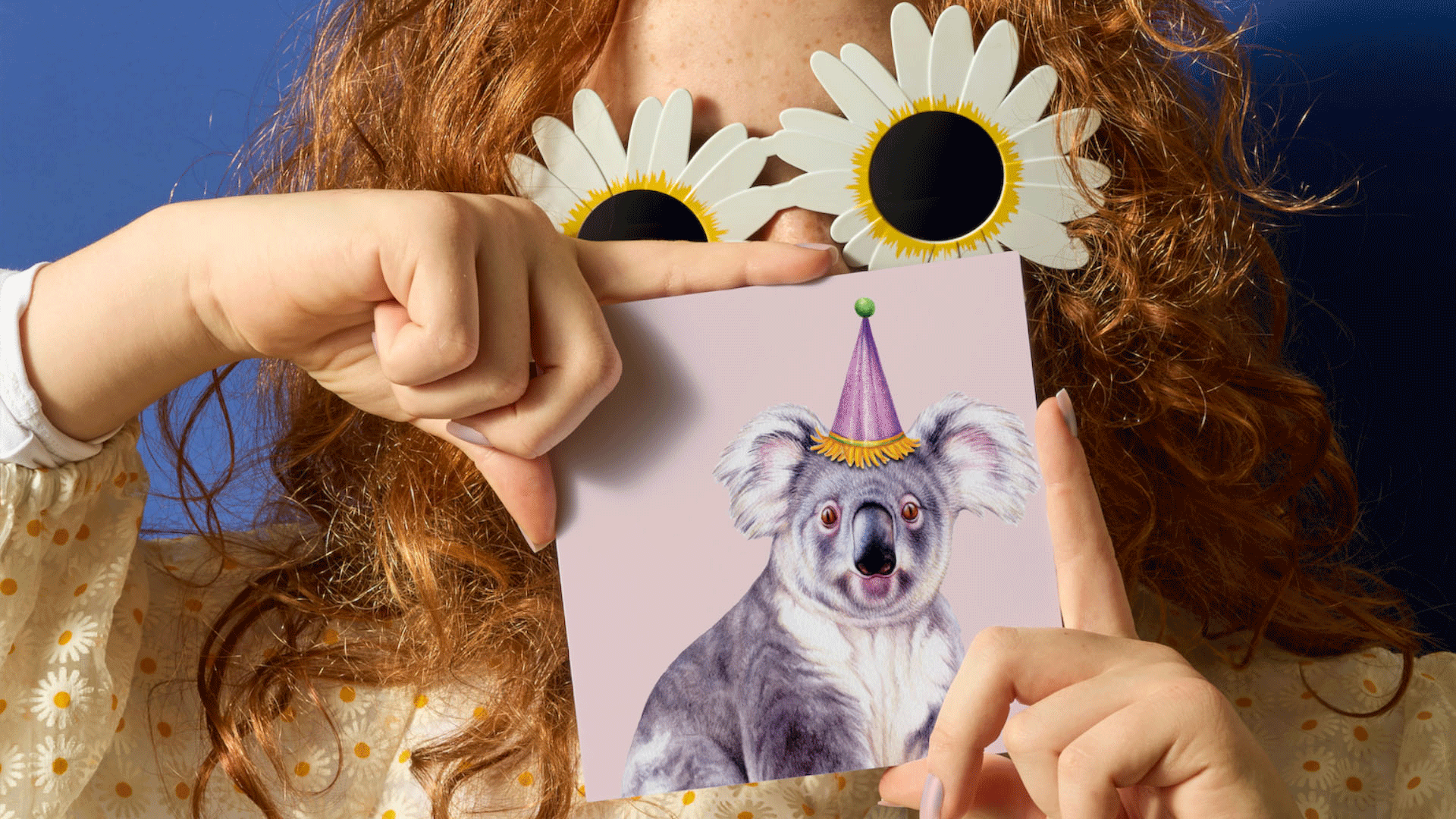 A person with curly red hair holds a square card featuring a koala wearing a party hat against a pink background. The person is wearing white daisy-shaped sunglasses and a cream floral top, posed against a dark blue background.